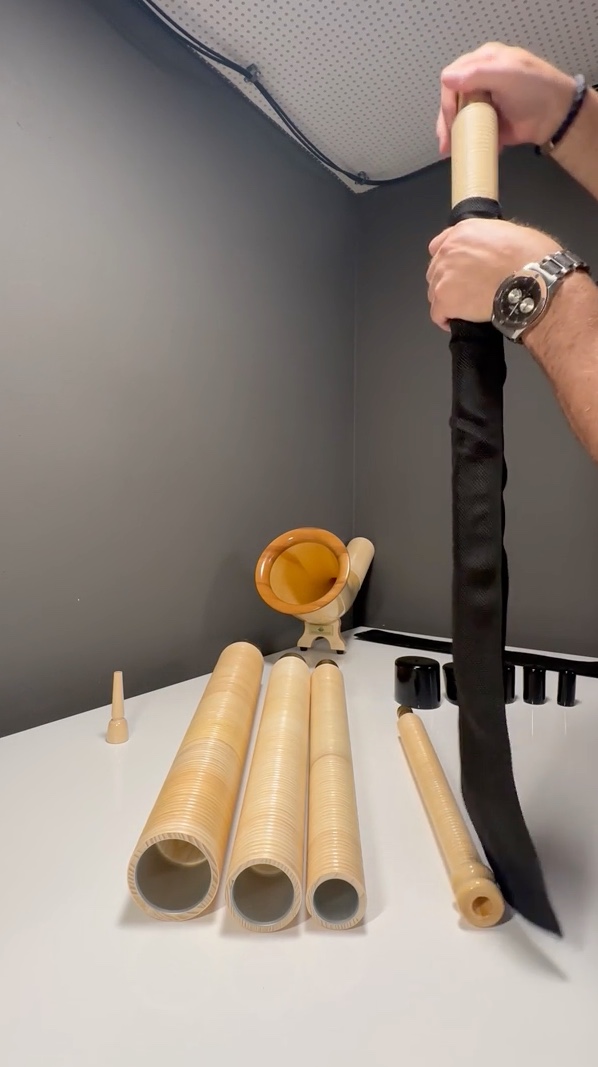 Montage eines Alphorns mit Holzteilen auf weißem Tisch, umgeben von Mundstücken und schwarzem Abdeckstoff.