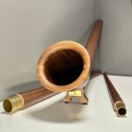 Alpenhorn auf Holzständer mit goldfarbenen Mundstücken, präsentiert auf glänzendem Tisch vor grauem Hintergrund.