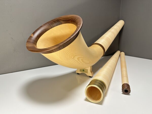 Alphorn mit Holzmundstück und Zubehör auf weißem Tisch vor grauer Wand.